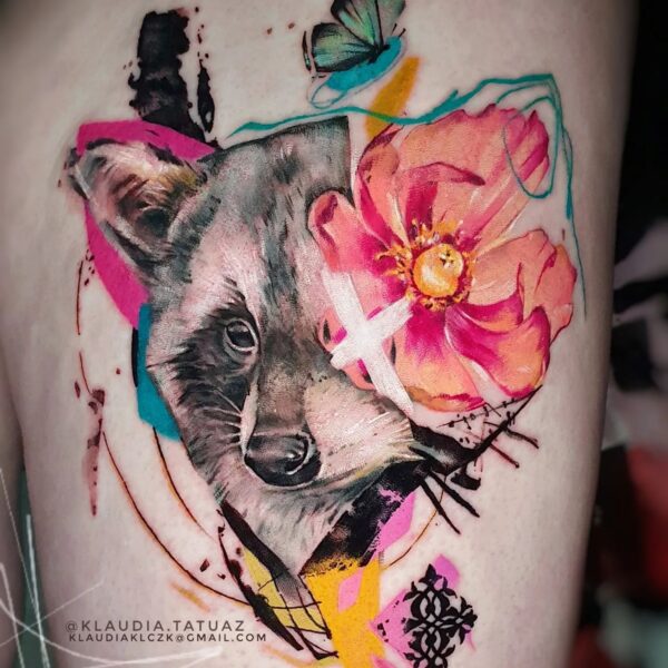 Abstract realism raccoon portrait tattoo on thigh with large pink flower and graphic colour splashes. Wildlife art tattooed at Silent Moon Tattoo Studio London UK.