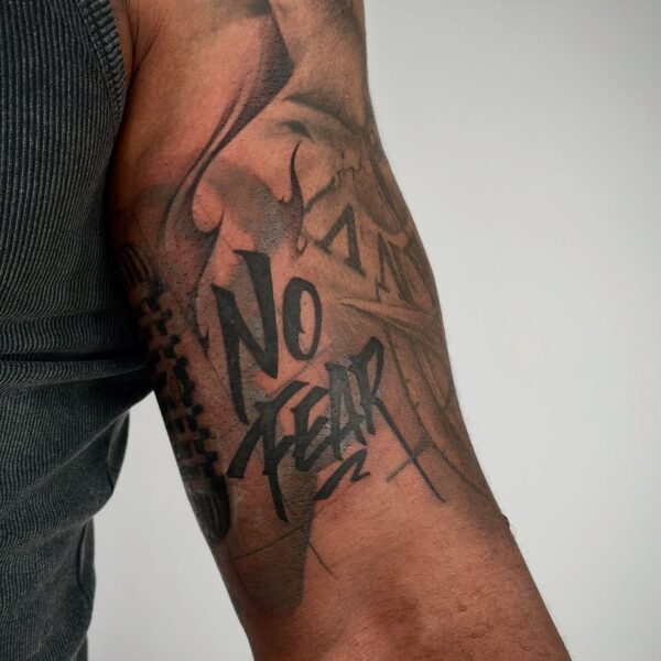 Bold blackwork script tattoo "No Fear" on the bicep in a brushstroke graffiti style, surrounded by abstract black and grey shading. Custom lettering tattooed at Silent Moon Tattoo Studio London UK.