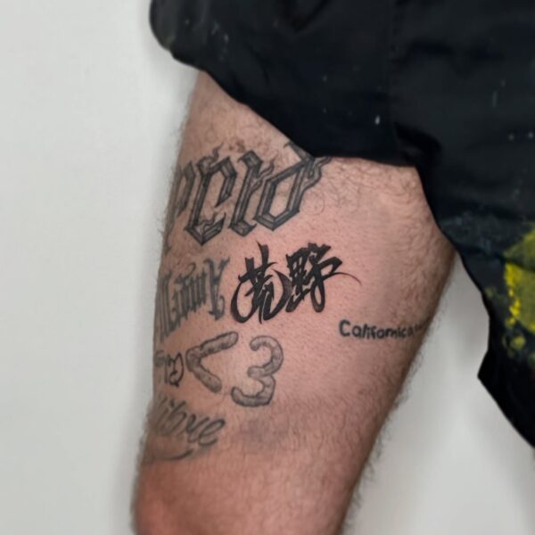 Blackwork calligraphy script tattoo in Old English and Japanese kanji for 'Wilderness' on the thigh. Custom lettering tattooed at Silent Moon Tattoo Studio London UK.