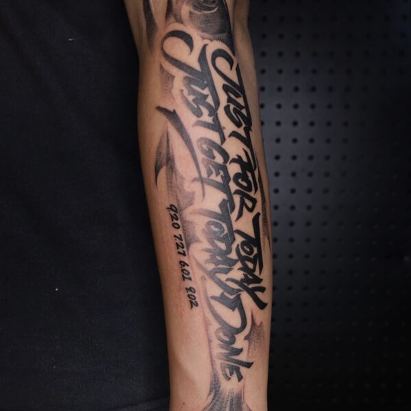 Custom calligraphy script tattoo "Just For Today Just Get Today Done" over a black and grey fish on the forearm. Blackwork lettering tattooed at Silent Moon Tattoo Studio London UK.
