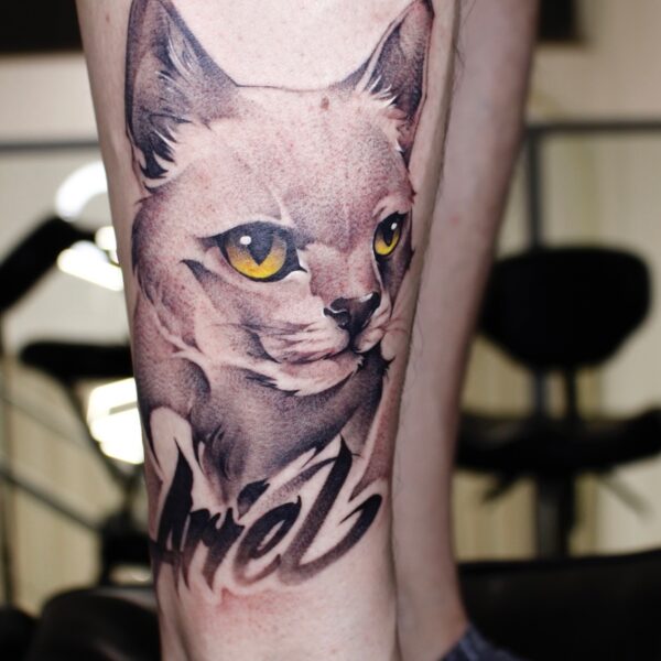 Colour realism cat portrait tattoo on the leg with bright yellow eyes and custom script name "Ariel" below. Pet memorial tattoo by Silent Moon Tattoo Studio London UK.