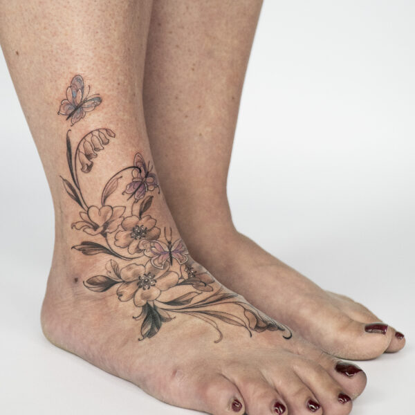 Delicate floral and butterfly ankle tattoo in soft colours by tattoo artist at Silent Moon Tattoo Studio in London.