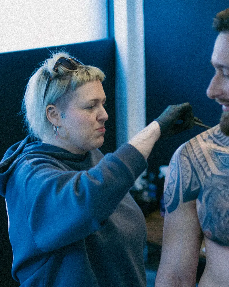 A candid, behind-the-scenes photograph of Klaudia, the highly experienced veteran tattoo artist and owner of Silent Moon Tattoo (SE16 London), providing precise care or preparation for a client's intricate, large-scale blue blackwork geometric tattoo pattern on his shoulder and chest, under cool blue studio lighting.