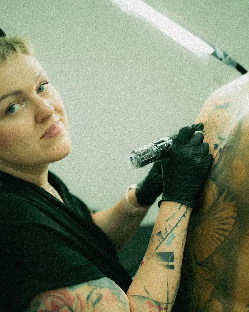 A close-up portrait of Klaudia, the highly experienced veteran tattoo artist and owner of Silent Moon Tattoo in London (SE16), looking at the camera with a gentle smile while actively tattooing a large-scale custom color piece on a client's back. She is wearing a black shirt and black nitrile gloves, holding a wrapped tattoo machine under bright studio lighting. Her own vibrant arm tattoos are clearly visible.