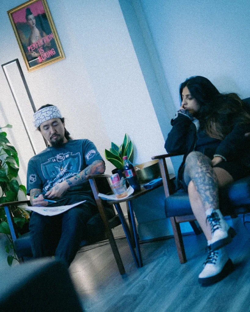 A candid, wide-angle photograph of experienced traditional tattoo artist Daisuke Sakaguchi during a bespoke design consultation in his London studio. Daisuke is seated on the left, wearing a blue t-shirt and a white patterned tenugui headband, focused on sketching a custom design on paper. On the right, a female client with visible leg tattoos sits in a mid-century modern chair, observing the process thoughtfully. The studio environment features a "Perfection is Boring" framed print, a snake plant, and a relaxed, professional atmosphere.