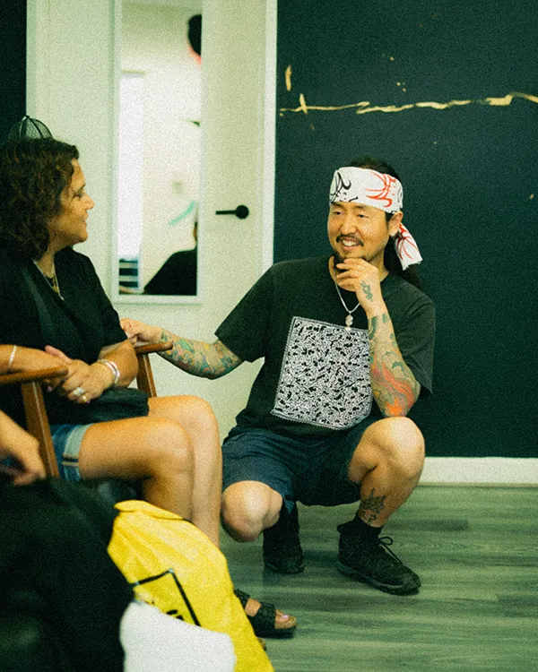 A candid, eye-level photograph of experienced traditional tattoo artist Daisuke Sakaguchi engaged in a mindful consultation with a client in his London studio. Daisuke is crouching in a relaxed, attentive posture, wearing a black graphic t-shirt and a white tenugui headband with red and black traditional patterns. His arms are covered in vibrant, multi-colored Irezumi sleeve tattoos. He is smiling warmly while listening to a seated client, illustrating his collaborative and bespoke approach to tattoo design.