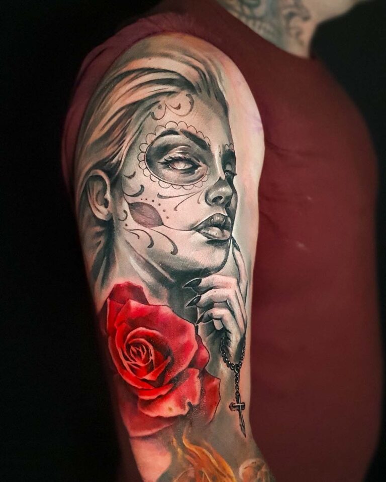 Day of the Dead girl tattoo on the upper arm, a woman's portrait with sugar skull face paint holding a rosary beside a red rose