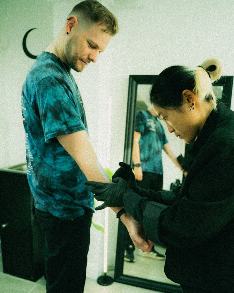 Behind the scenes at Silent Moon, tattoo artist Satrxx placing a stencil on a client's inner forearm while the client stands to check the position, with a mirror and the studio's crescent moon on the wall